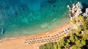 Vista aérea de una playa con aguas turquesas cristalinas, una fila de tumbonas y un afloramiento rocoso.