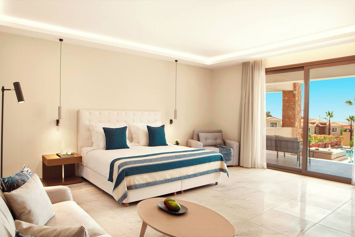 Habitación de hotel moderna con una cama grande, un sofá, una mesa de centro y vistas a la piscina y al área exterior.