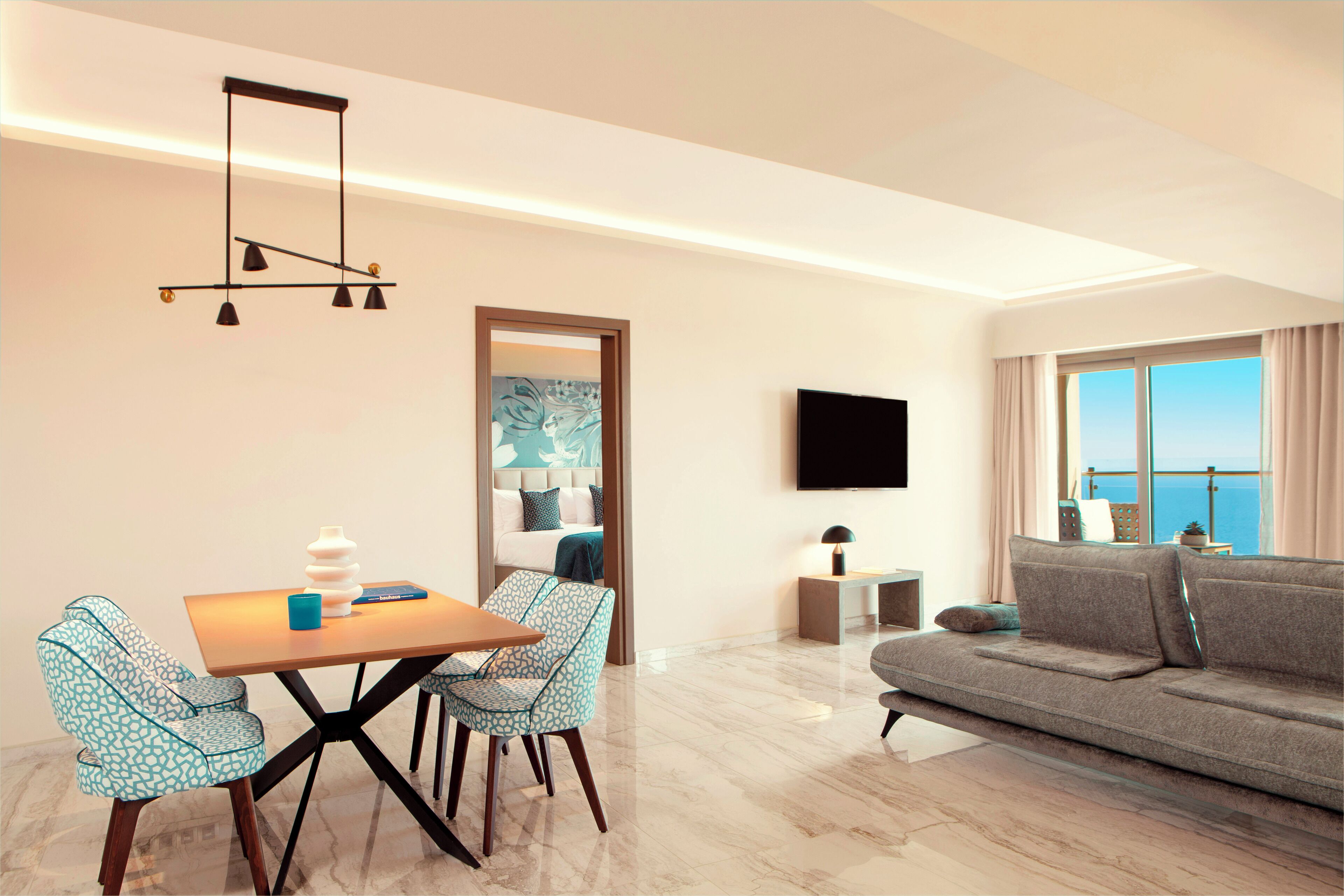 A modern living room with a sofa, a dining table with chairs, a TV mounted on the wall, and a large window with a view.