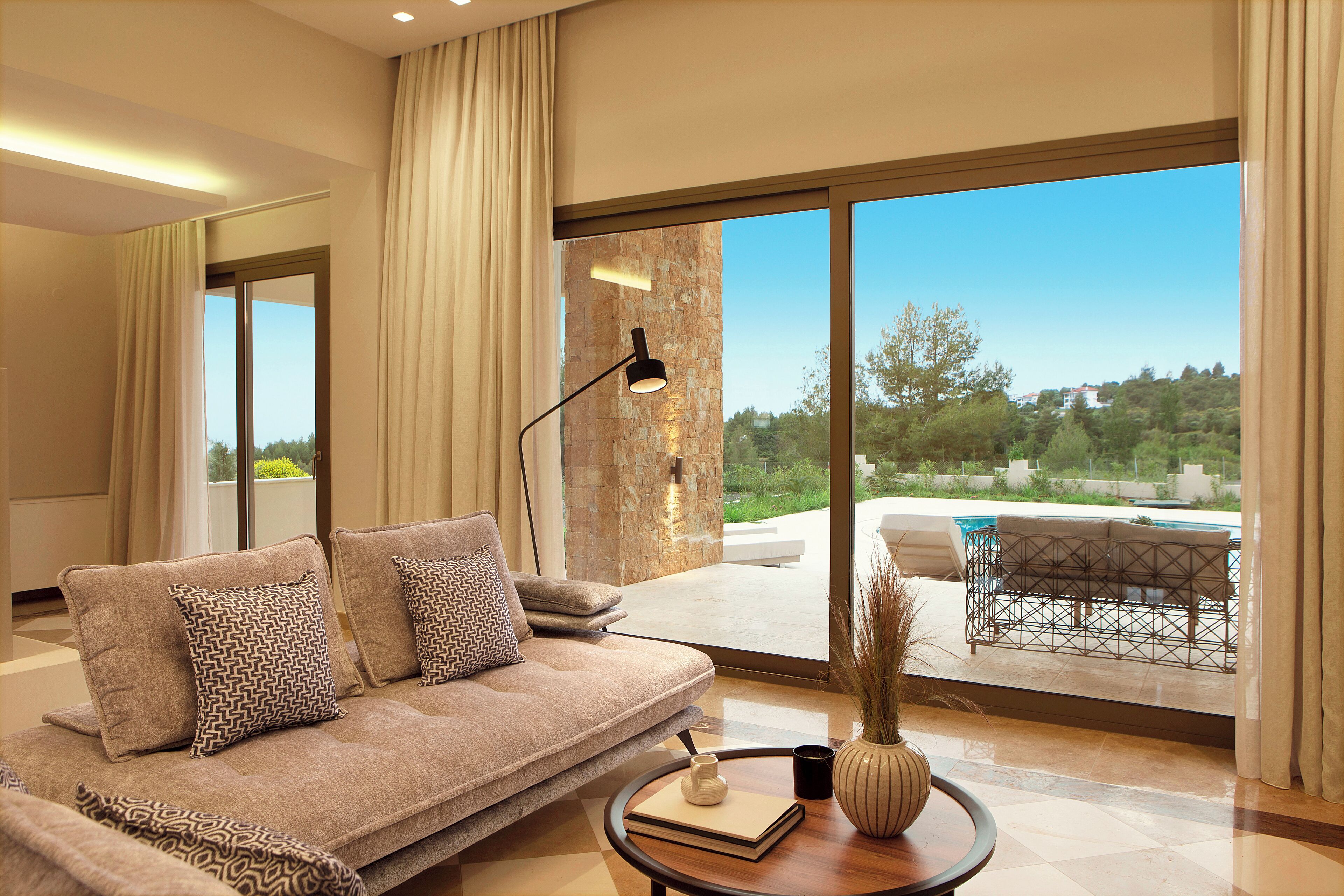 A modern living room with a sofa, a coffee table, and a view of a swimming pool and greenery.