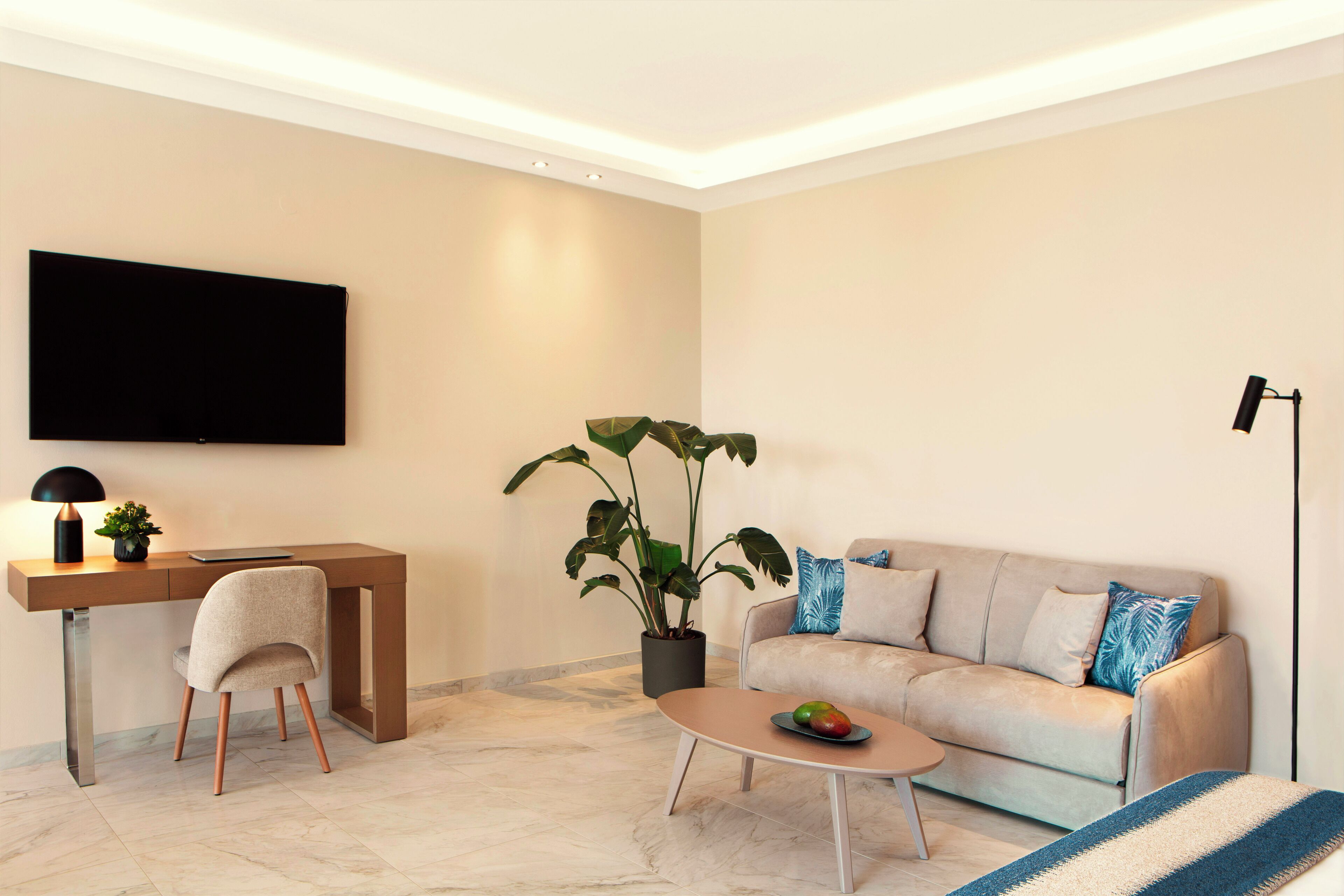 A modern living room with a flat-screen TV, a desk, a comfortable sofa, a coffee table with fruit, and a potted plant.