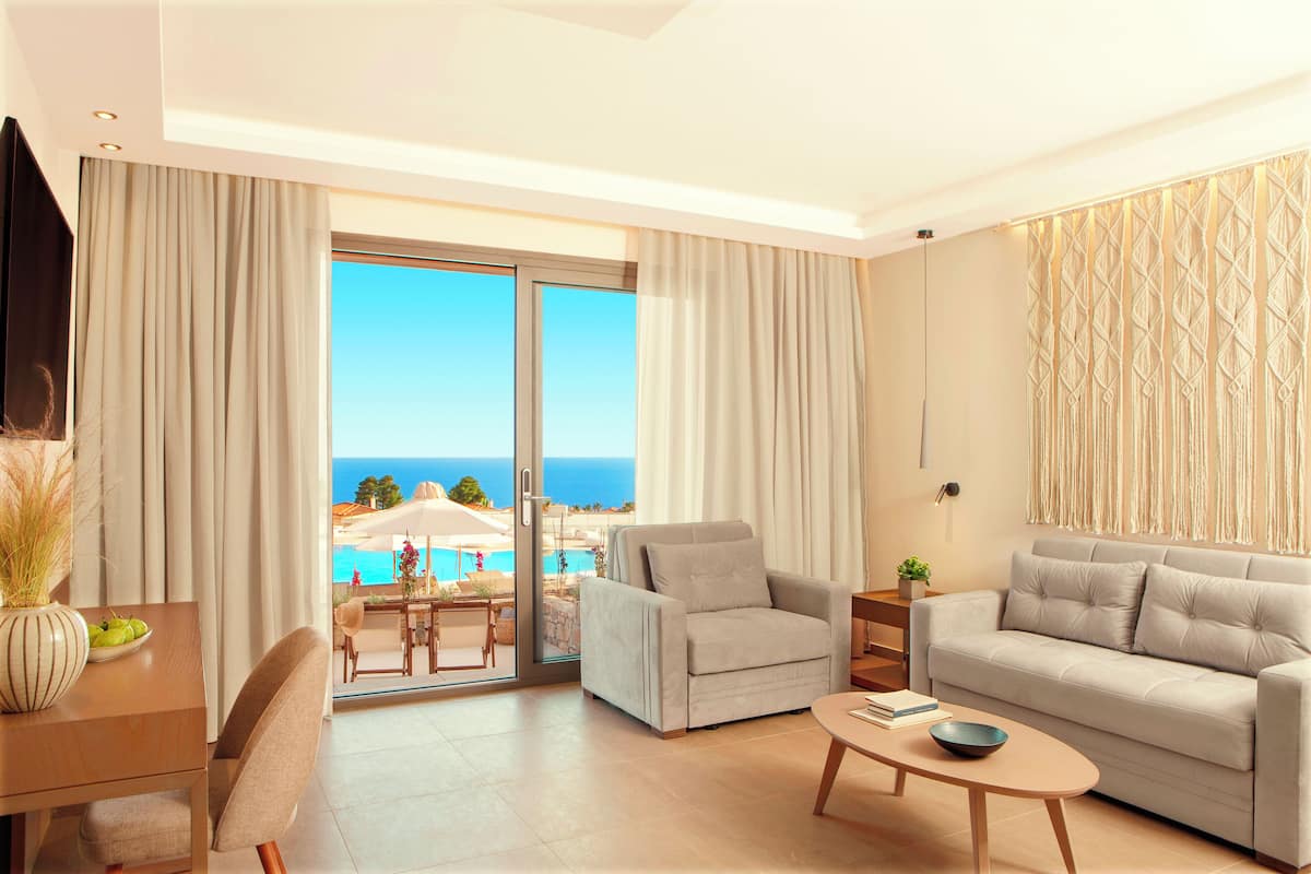 Una sala de estar moderna con sofá, sillón y mesa de centro. Un amplio ventanal corredera da a un balcón con vistas al océano y a una piscina.