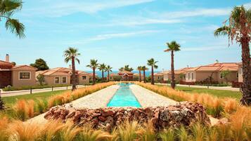 Una piscina con borde de piedra, rodeada de césped y palmeras, que da a una hilera de casas con tejados rojos.