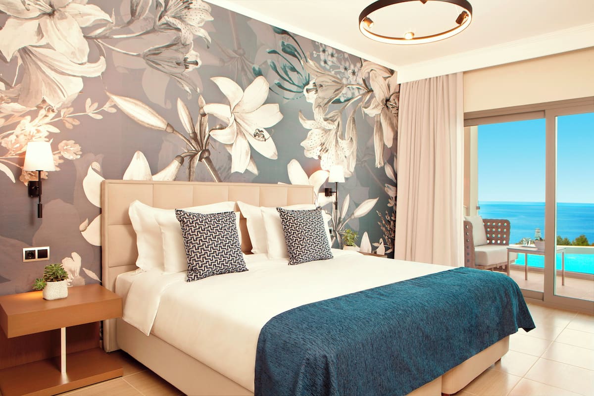Un dormitorio con una cama grande, papel tapiz floral, vista al mar y un balcón con mesa y sillas.