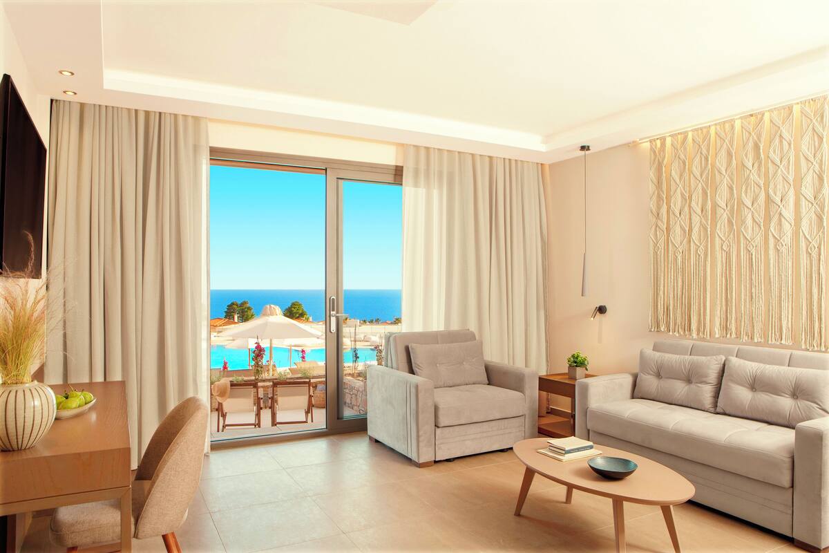 Una sala de estar moderna con un sofá, sillones y una mesa de centro. Un amplio ventanal corredero da a un balcón con vistas al océano y una piscina.