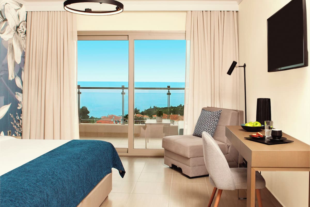 Habitación de hotel con cama, escritorio, silla, televisor y balcón con vista al mar.