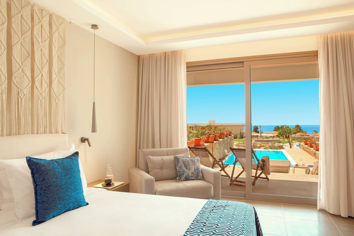 Habitación de hotel con cama, sofá, vista a la piscina y al mar, y balcón con mobiliario exterior.