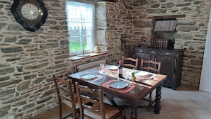 Comedor de paredes de piedra con una mesa de madera dispuesta para cuatro, un reloj de pared grande y un ventanal con vista a un jardín.