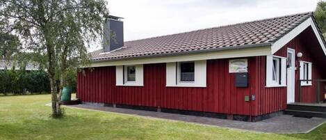 A red building with a tiled roof, a small porch, and a green trash bin.