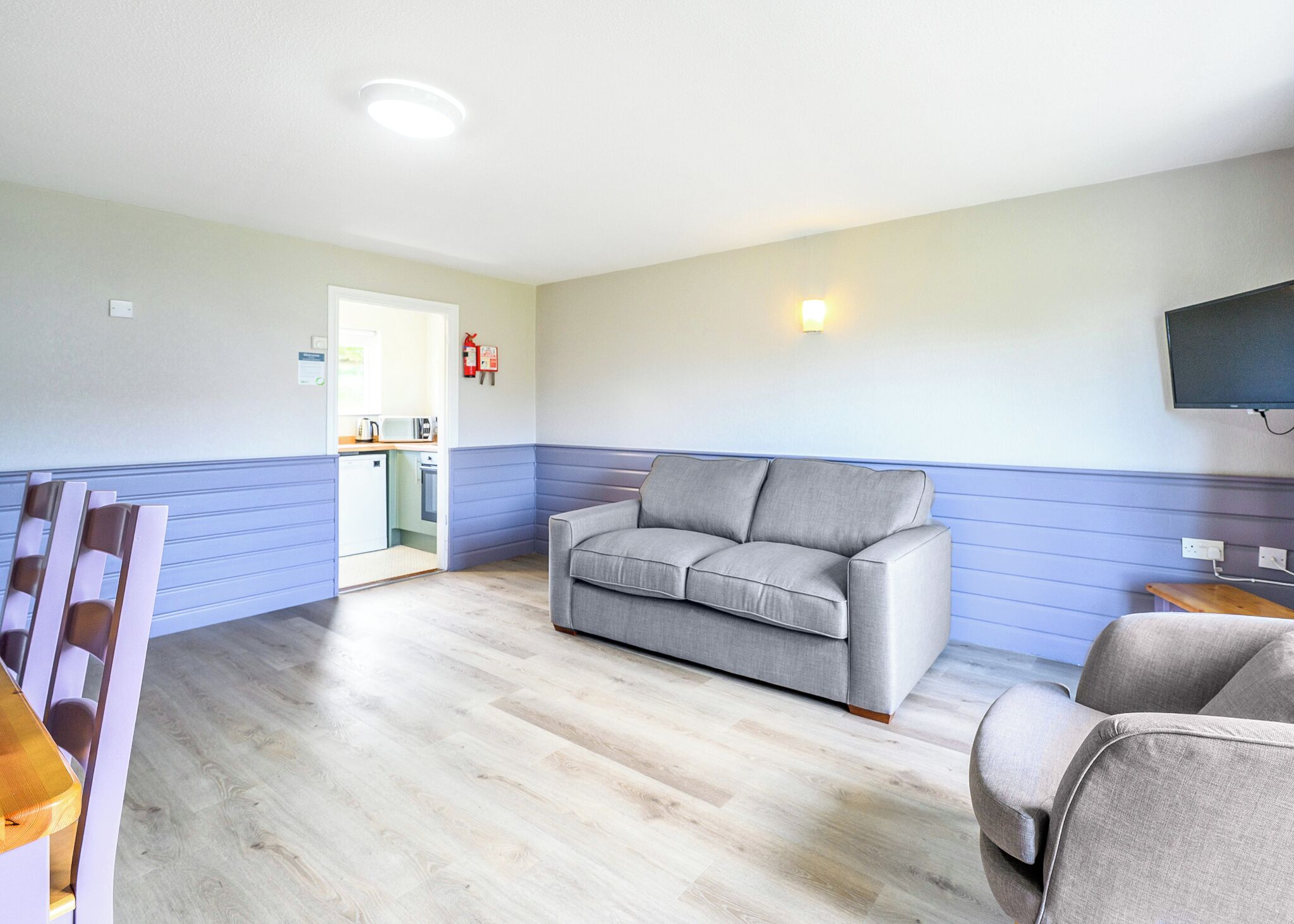 A room with a grey sofa, a wooden table, a television mounted on the wall, and a kitchen area visible through an open door.
