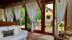 A cozy room with a sofa, a chair, and a table. There are large windows with curtains, and a view of a garden and a building outside. - Chalet in Styria with Pool & Panoramic Views (Fehring)