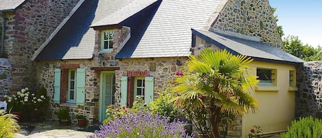 A stone building with a sloped roof, a small garden with lavender, and a stone pathway.