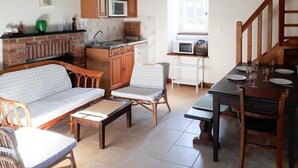 A cozy living room with a sofa, chairs, a coffee table, a dining table set for two, and a kitchen area with wooden cabinets and a brick fireplace. - Stone House in Plurien near Sandy Beaches (Plurien)