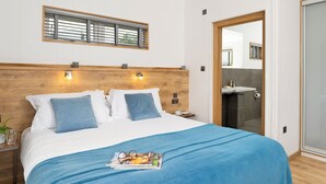 Ein Hotelzimmer mit einem hölzernen Kopfteil, einem großen Fenster mit Jalousien, einem Badezimmer mit Spiegel und Waschbecken sowie einer blauen Bettdecke.