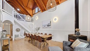 A spacious living area with a high ceiling, wooden beams, a dining table with chairs, a sofa, a fireplace, and a staircase leading to an upper level.