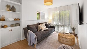 A living room with a grey sofa, white shelving, a woven coffee table, and a large window with sheer curtains.