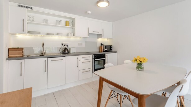 Ett modernt kök med vita skåp, en inbyggd ugn och en matplats med ett vitt bord och stolar.