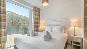 Ett hotellrum med en stor säng, två sänglampor, en träpanel vid sänggaveln, en balkong med utsikt över träd och kullar samt ett fönster med gardiner.