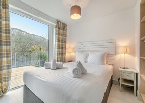 Habitación de hotel con una cama grande, dos lámparas de noche, un cabecero de madera, un balcón con vistas a árboles y colinas, y una ventana con cortinas.