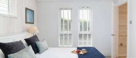 A bedroom with a bed, pillows, a tray with a mug and fruit, and a view through white shutters.