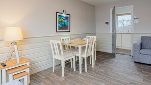 Un coin repas comprenant une table et des chaises, une lampe, un tableau encadré, et donnant sur une fenêtre et une porte.