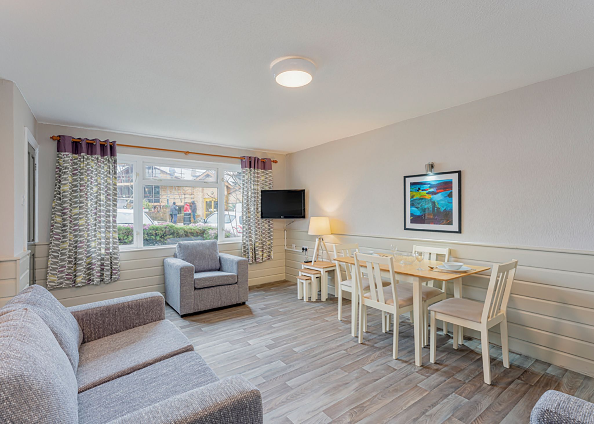 Una habitación con sofá, una mesa de comedor con sillas, un televisor montado en la pared y una ventana con cortinas.