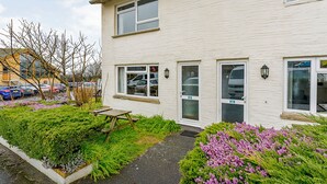 Un bâtiment à deux étages, à la façade blanche, avec un petit jardin agrémenté de fleurs violettes et une table de pique-nique.