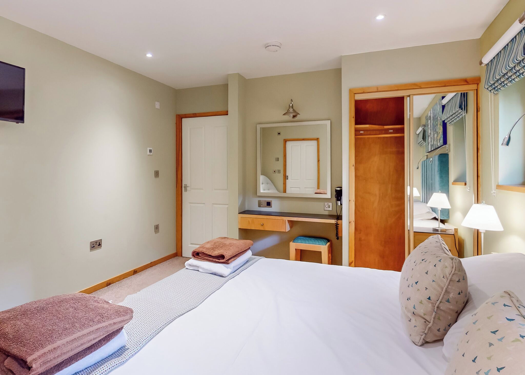 Quarto de hotel com cama, escrivaninha, espelho, guarda-roupa e banheiro.