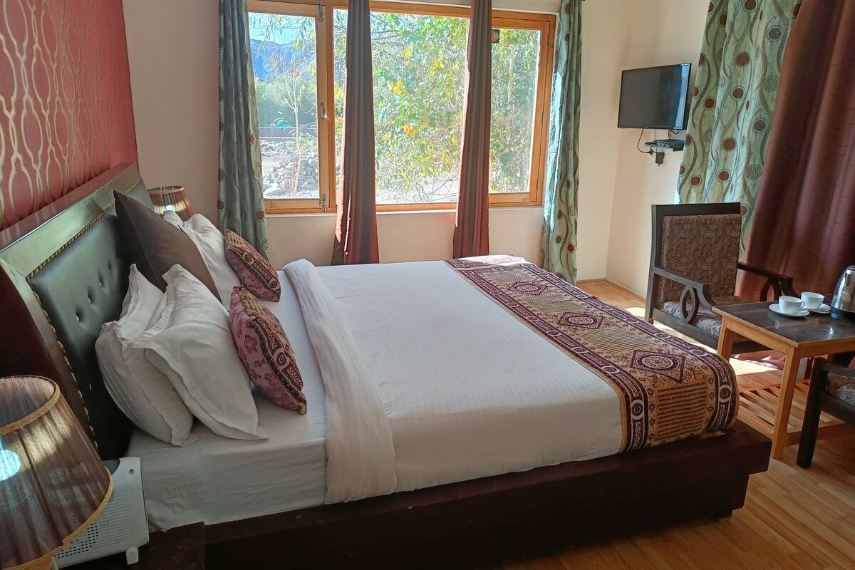 A neatly made bed with a patterned bedspread, a wooden nightstand with a lamp, a chair, and a table with cups and a kettle.