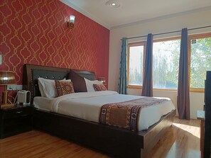 A bedroom with a large bed, a nightstand with a lamp, a wall-mounted light fixture, and a window with curtains. - Munna Palace  (Leh)