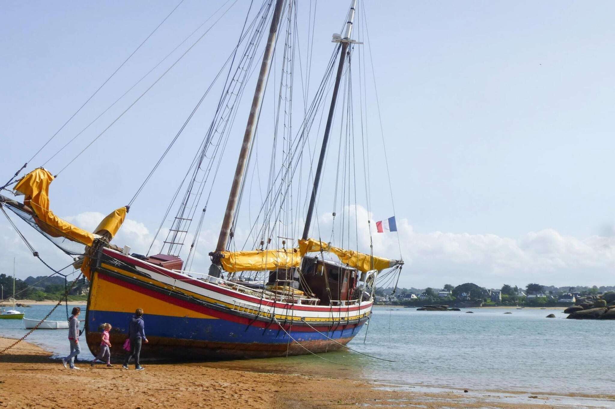Een kleurrijke boot met masten en zeilen, afgemeerd aan een zandstrand met een rustige zee en gebouwen in de verte.