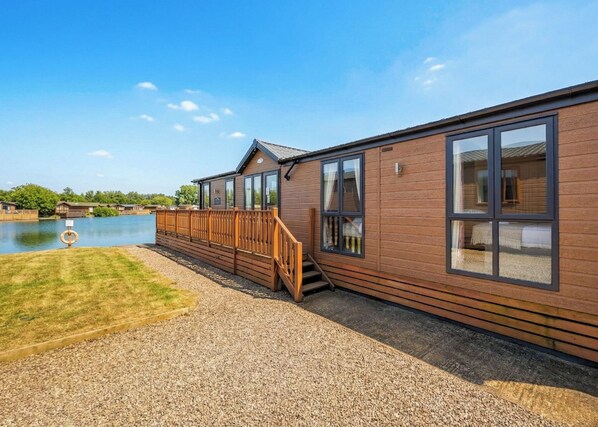 Un bâtiment en bois doté de grandes fenêtres, d’une terrasse et d’un chemin de gravier menant à un plan d’eau.