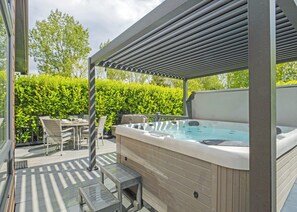 Un jacuzzi bajo una pérgola, rodeado por una terraza de madera y mobiliario exterior.