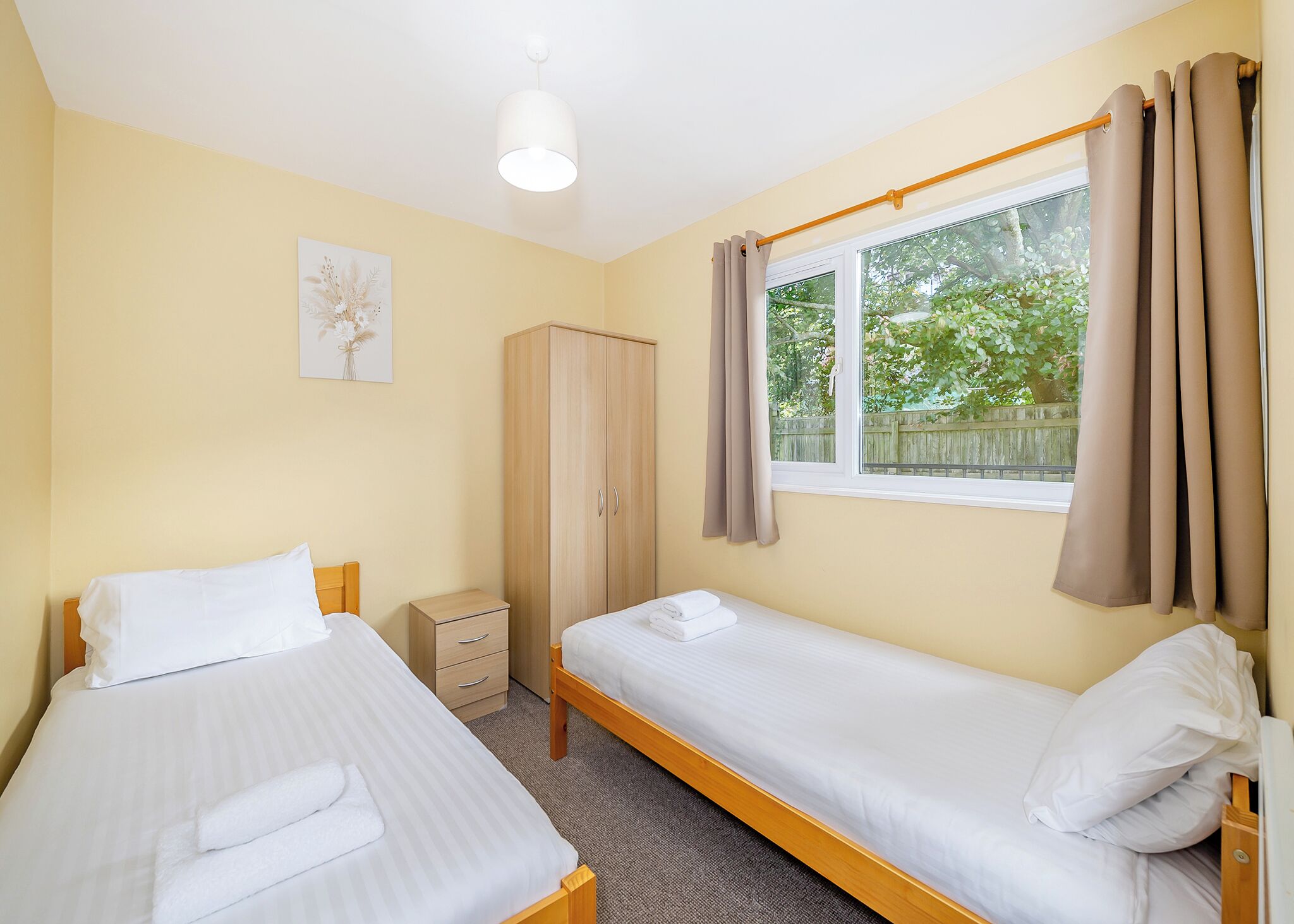 Habitación con dos camas individuales, un armario, una mesita de noche pequeña, una ventana con cortinas y una lámpara en el techo.