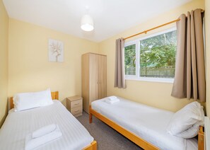Ein Zimmer mit zwei Einzelbetten, einem Kleiderschrank, einem kleinen Nachttisch, einem Fenster mit Vorhängen und einer Deckenleuchte.