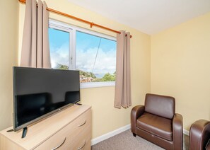 A room with a TV on a wooden dresser, a brown leather chair, a window with curtains, and a view of buildings and greenery.