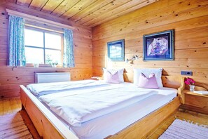 A wooden cabin bedroom with a double bed, a window with curtains, a bedside table with a lamp, and a framed picture on the wall.