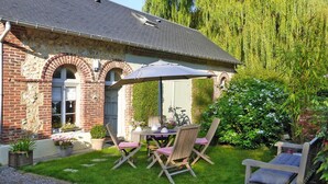 Een bakstenen huis met een patio tafel en stoelen, omringd door groen en een treurwilg.