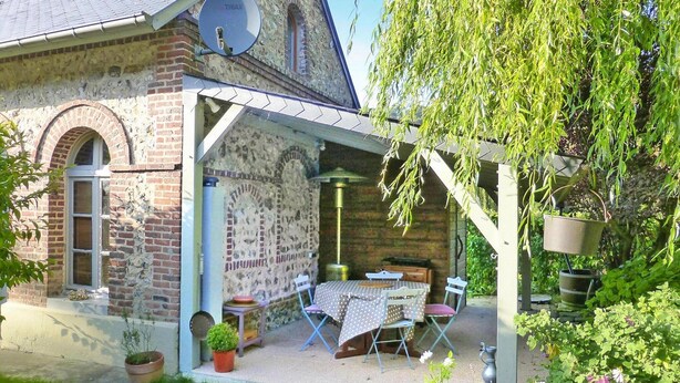 Een landelijk stenen gebouw met een overdekt terras, een tafel met stoelen en potplanten.