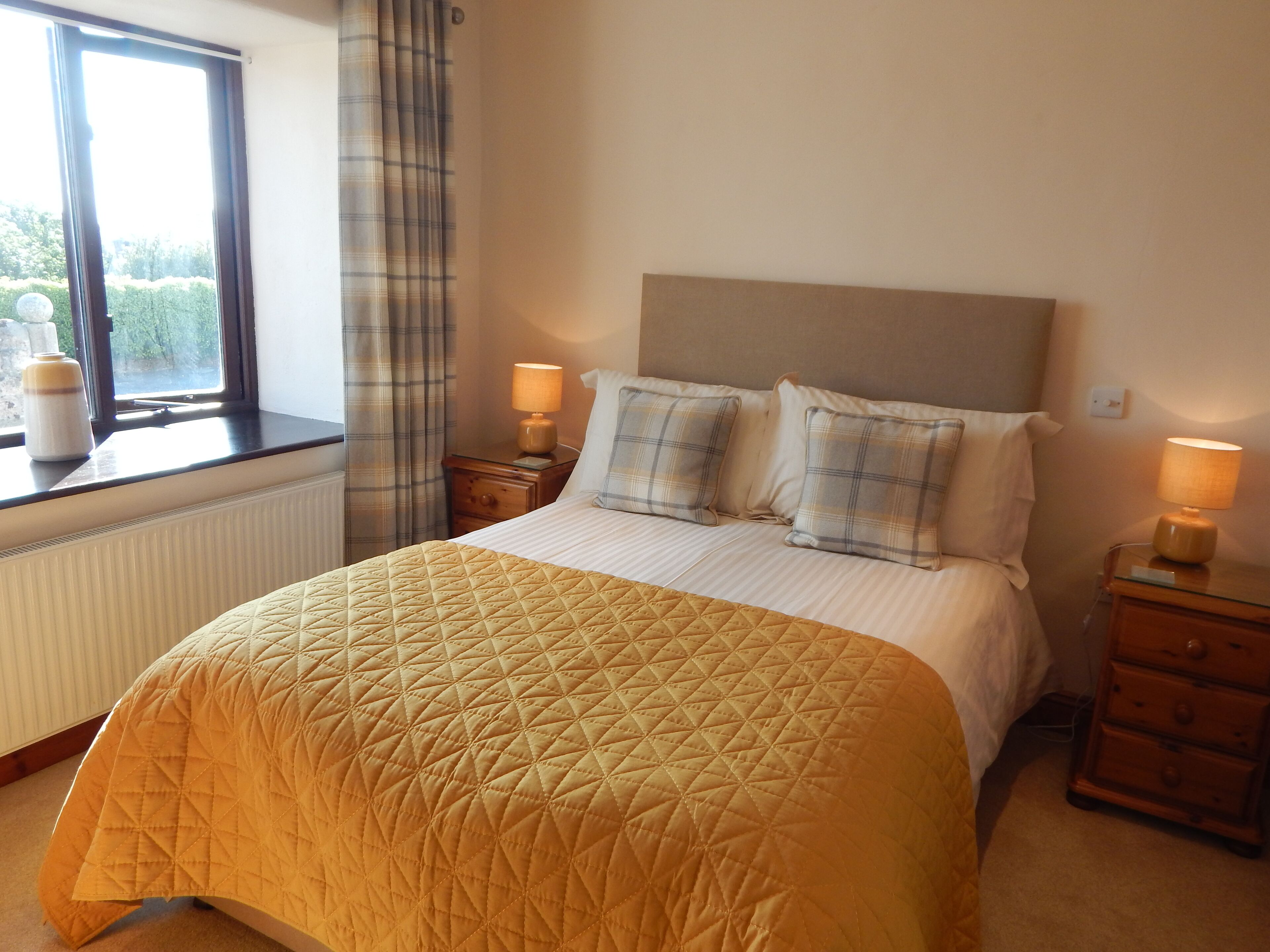 A neatly made bed with a quilted cover, two bedside lamps, a window with curtains, and a radiator.