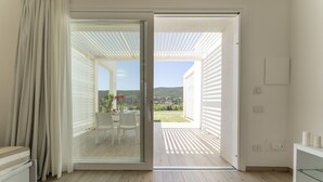 Puerta corrediza de vidrio abierta que da a un patio exterior con mesa y sillas, vista a una colina, y un interior blanco con un armario de cristal.