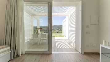 An open sliding glass door leading to an outdoor patio with a table and chairs, a view of a hillside, and a white interior with a glass cabinet.