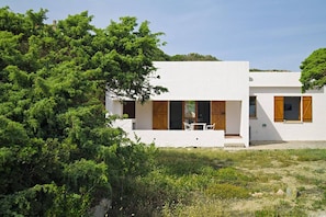 A modern white building with wooden shutters, a patio with outdoor seating, and lush greenery surrounding it.