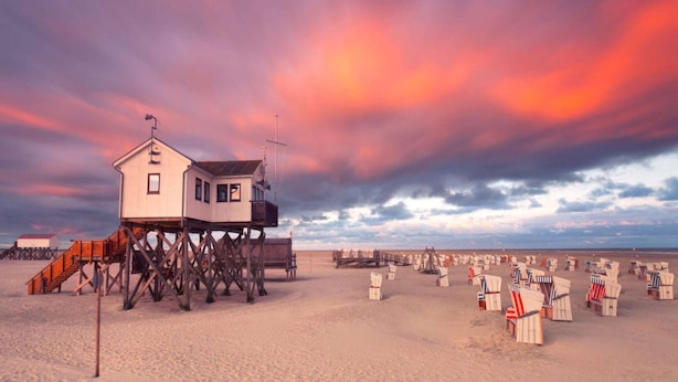 Une maison de plage sur pilotis, avec un escalier, des chaises de plage et des parasols.