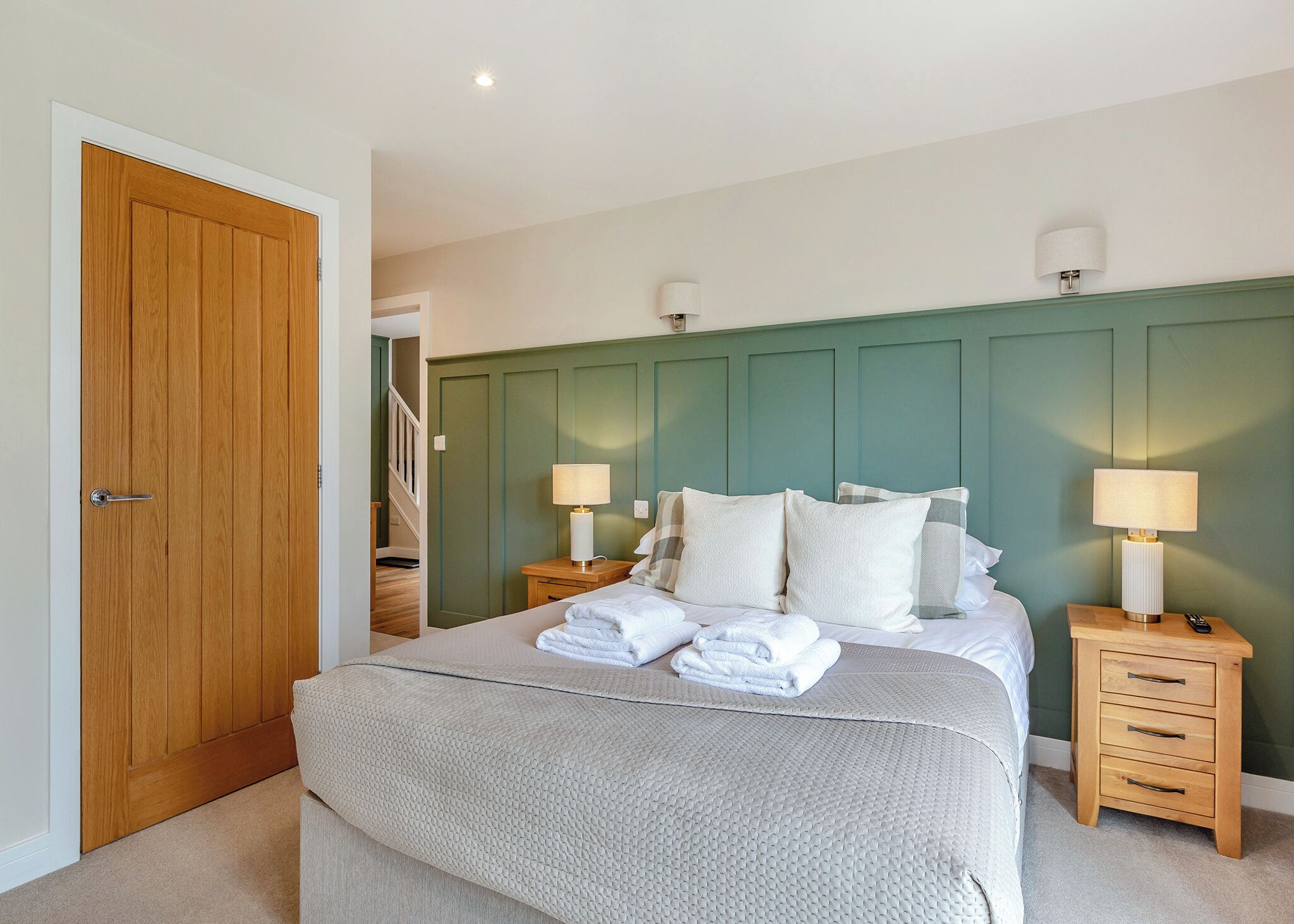 Un dormitorio con puerta de madera, una cama con ropa de cama blanca, dos mesitas de noche con lámparas y un cabecero verde.
