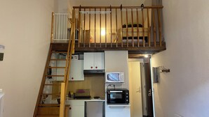 Een compact appartement met een kitchenette, een kleine koelkast, een magnetron, en een slaapverdieping van hout met een balkon.