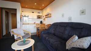 A living room with a dark sofa, a small round table, and a wicker chair. There is a kitchen in the background with white cabinets and a wooden ceiling. - Ferienwohnung Tiefblick, Dornumersiel (Dornumersiel)