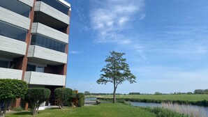 A modern building with multiple balconies, a grassy area with a tree, and a river in the background. - Ferienwohnung Tiefblick, Dornumersiel (Dornumersiel)