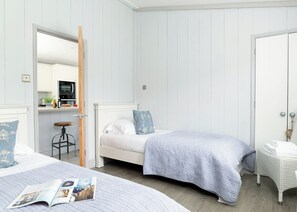 Une chambre avec deux lits, un petit tabouret, une chaise en osier et une porte donnant sur la cuisine.