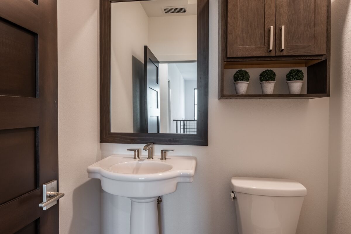A bathroom with a white sink, a mirror, and a toilet. There are three potted plants on a shelf above the toilet.
